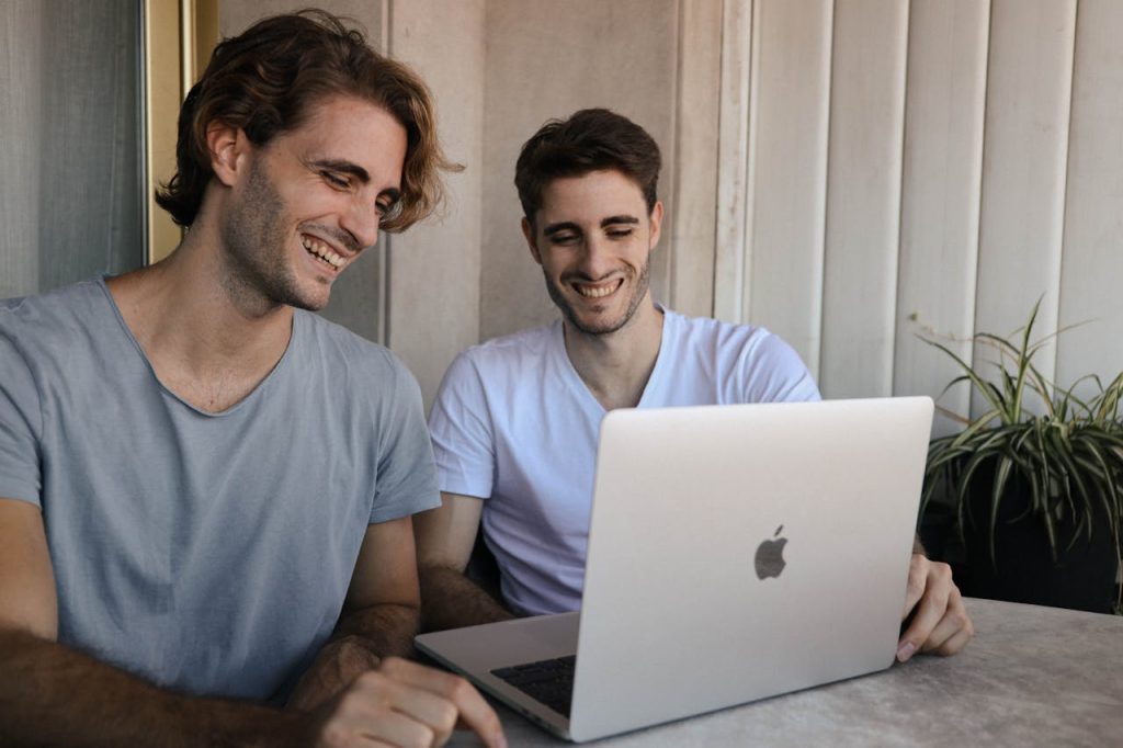 Two young men laughing while working together on a laptop outdoors, showcasing teamwork and friendship.