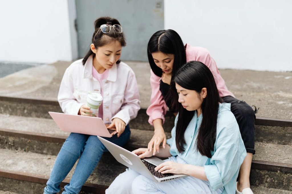 Three women sitting on stairs working together on laptops, enjoying teamwork outdoors.