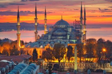 On a sunset atmosphere, an old mosque, surrounded by smaller building structures and a few trees.