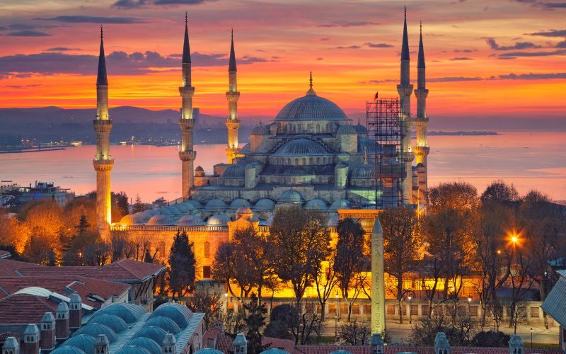On a sunset atmosphere, an old mosque, surrounded by smaller building structures and a few trees.